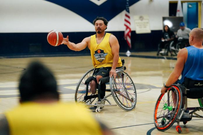 A male wheelchair basketball player wearing a yellow jersey throws a ball during a game